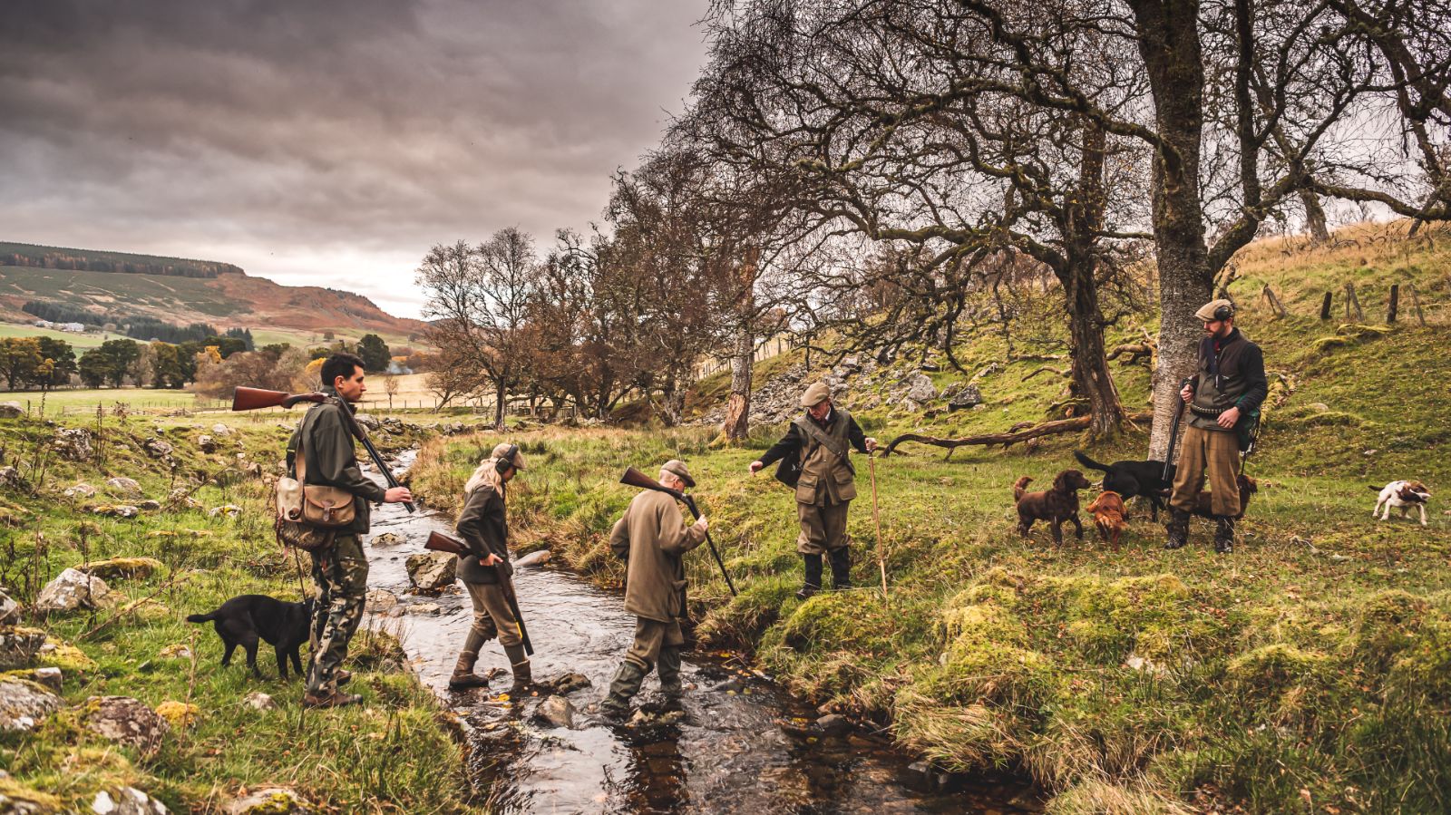 group of people and their dogs crossing river, wearing wellies and carrying shotguns and cartridge bags