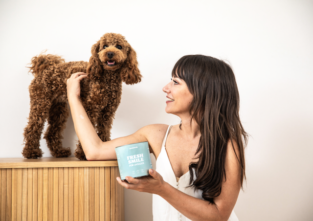 brown haired white woman holding a tub of mammaly treats and looking at her dog - a brown poodle - which is on her left